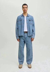 Denimo striukė su dviem krūtinės kišenėmis ir priekiniais mygtukais, dėvima ant baltos marškinėlių su siaura apykakle, derinama su laisvai krentančiais šviesiai mėlynais džinsais ir béžiniais batais.