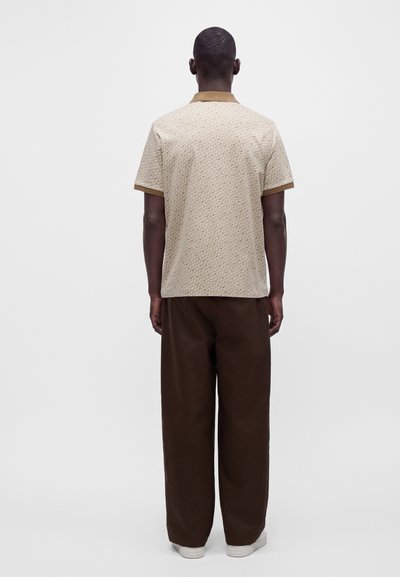 Homme portant un polo beige à manches courtes avec bordure marron et un pantalon large marron foncé, debout, dos tourné sur un fond uni.