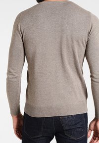 Beige stickad tröja med långa ärmar, ribbade manschetter och nederkant, har rund halsringning och en slät yta, sedd bakifrån.