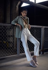 Light green blazer over a beige top, paired with white straight-leg pants and gray sneakers, against a metal fence in an industrial setting.