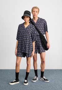 Dos personas visten camisetas de manga corta negras a juego y pantalones cortos con un patrón de hojas moradas. Una de ellas lleva un sombrero tipo cubo de rayas negras y lleva una bolsa texturizada.