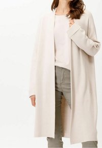 Jasno beżowy długi kardigan wykonany z miękkiej tkaniny, z otwartym przodem, szerokimi rękawami i dwoma bocznymi kieszeniami, zestawiony z jasnym topem i zielonymi spodniami.