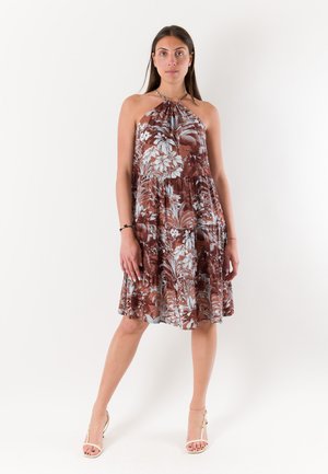 Donna in piedi con le gambe divaricate che indossa un vestito marrone floreale senza maniche con collo all'americana e sandali beige con tacco alto su sfondo bianco.