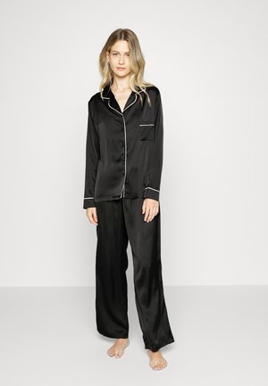 Vrouw staat op blote voeten en draagt een zwart satijnen pyjamajasje met knopen en lange mouwen, gecombineerd met bijpassende wijde pijpen op een effen witte achtergrond.