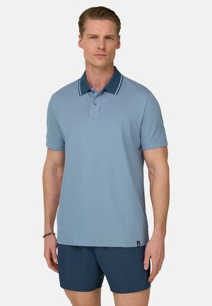 Mand iført en lyseblå polo med mørkeblå krave og mørkeblå shorts, stående mod en hvid baggrund.