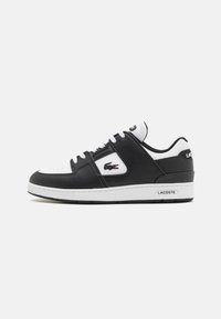 Zapatilla negra de cuero con acentos blancos, puntera redonda, cierre con cordones, cuerpo texturizado y logo de Lacoste en el lateral. Suela de goma.