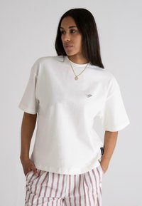 White cotton t-shirt with short sleeves, round neckline, small purple logo on the chest, and a relaxed fit. Paired with striped pants.