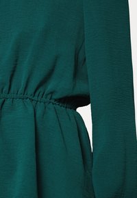 Blouse couleur sarcelle avec une taille élastique froncée, des manches longues et un tissu texturé à l'aspect légèrement froissé.