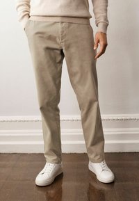 Beige Chinos mit glatter Textur, schmaler Passform und seitlichen Taschen, kombiniert mit weißen Turnschuhen auf einem Holzboden.