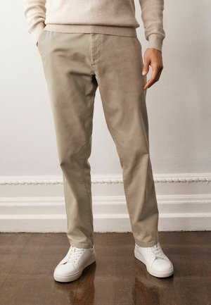 Beige chino's met een gladde textuur, tapered fit en zijzakken, gecombineerd met witte sneakers op een houten vloer.