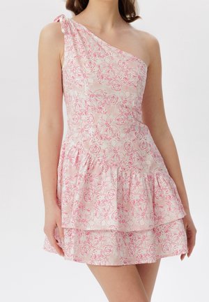 Kvinde iført en lyserød og hvid blomsteret one-shoulder kjole med flæsedetaljer og en sløjfe på skulderen, stående mod en ensfarvet baggrund.