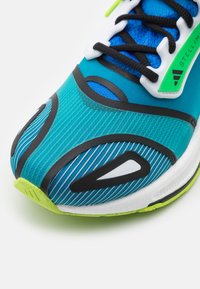 Tealové a modré sportovní boty se strukturovaným svrškem se pruhy, černými akcenty, bílými detaily a jasně zelenou podešví.