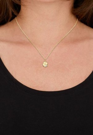 Collar de oro con un charm en forma de trébol de cuatro hojas, con un pequeño acento de gema, usado sobre una camisa negra.