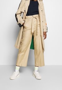 Beige Trenchcoat mit schwarzen Akzenten, lockere beige Hose mit Bindegürtel und weiße Sneakers mit grün- und gelbgestreiften Socken.