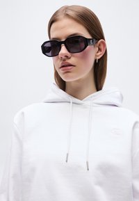 Młoda kobieta z prostymi brązowymi włosami, nosząca czarne, duże okulary przeciwsłoneczne, małe kolczyki koła oraz białą bluzę z kapturem i sznurkami.