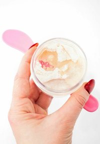 Klar glasbeholder med pulver og en lille mængde fløde. En pink plastikspartel hviler mod beholderen. En hånd, der holder beholderen, er synlig.