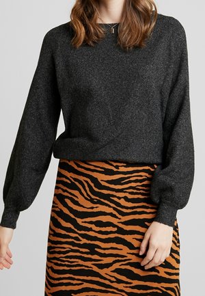 Frau trägt einen dunkelgrauen Pullover mit langen Ärmeln und einen tigerstreifenmusterierten orange-schwarzen Rock, steht vor einem einfarbigen Hintergrund.