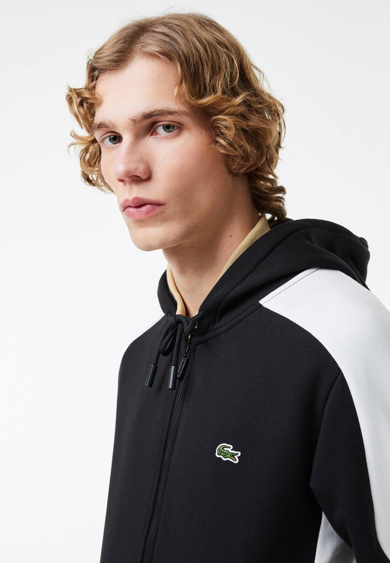 Sweat à capuche noir avec des manches accentuées en blanc, fermeture éclair à l'avant, cordon de serrage et logo crocodile brodé en vert sur la poitrine. Texture lisse.