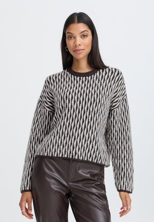 Vrouw met lang donker haar, die een zwart-wit gestreepte trui en donkerbruine leren broek draagt, staand tegen een effen lichte achtergrond.