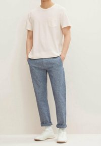 Mężczyzna w gładkiej, białej koszulce z krótkim rękawem, niebieskich spodniach do kostek, białych skarpetkach i białych trampkach, stoi z rękami w kieszeniach.