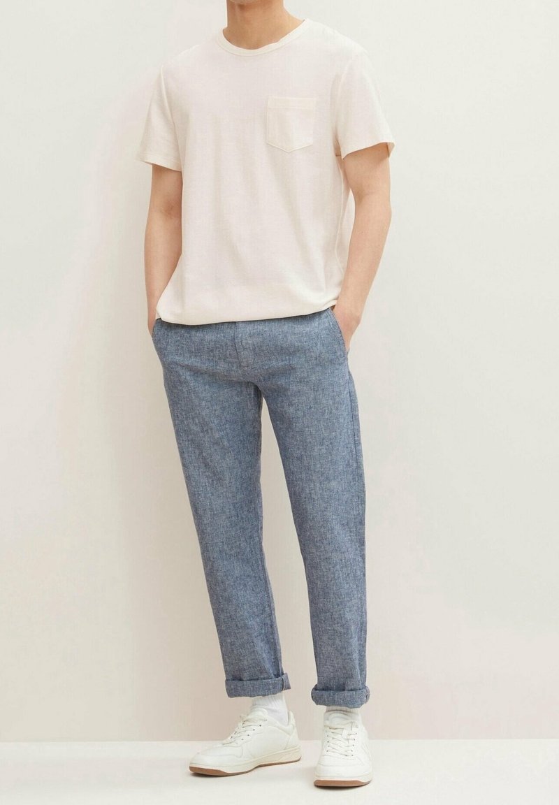 Mężczyzna w gładkiej, białej koszulce z krótkim rękawem, niebieskich spodniach do kostek, białych skarpetkach i białych trampkach, stoi z rękami w kieszeniach.