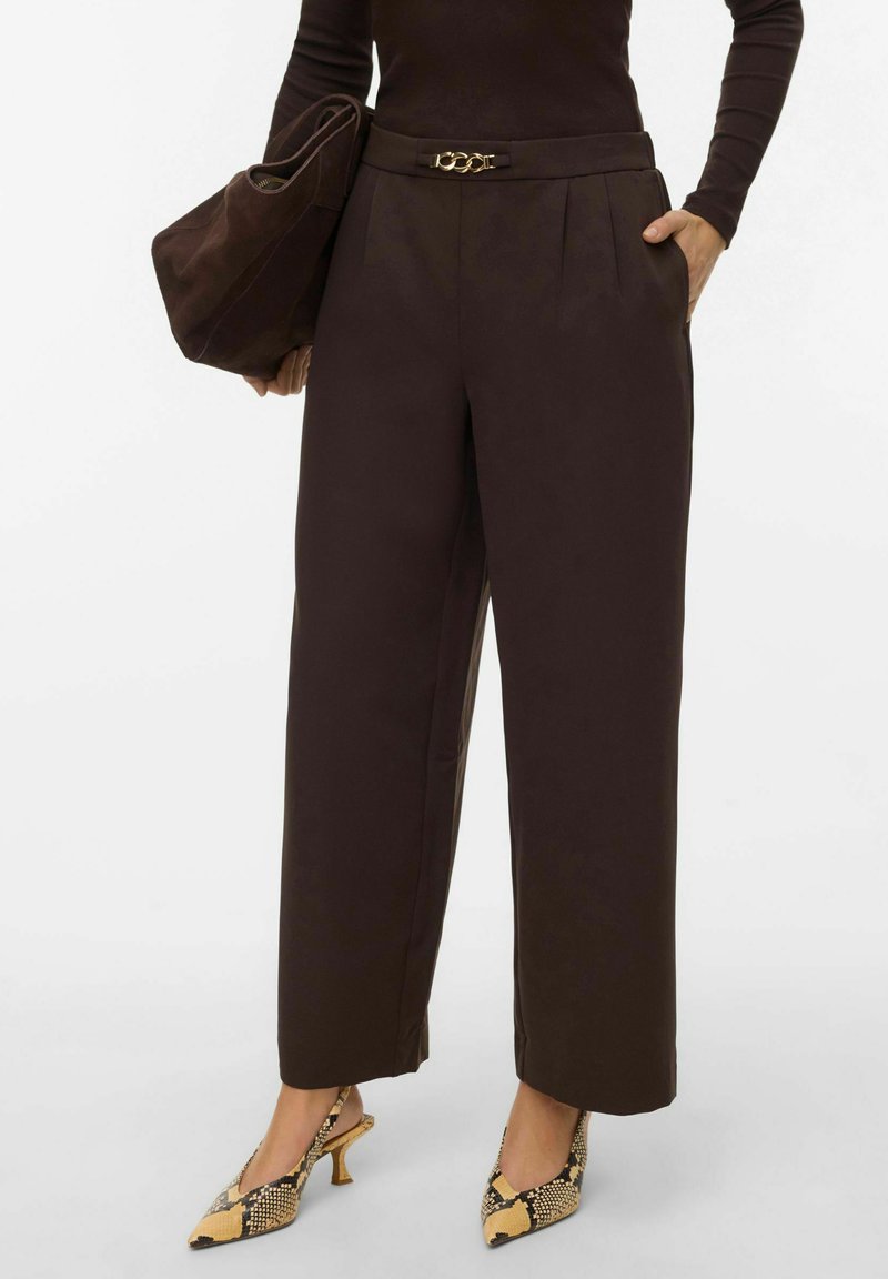Pantalon ample marron avec ceinture à chaîne dorée, associé à un sac marron et des chaussures à talons hauts à motif imprimé serpent.