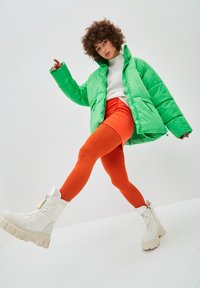Chaqueta acolchada verde con cuello alto, leggings naranjas y botas de plataforma blancas con acentos texturizados. Modelo posando de manera dinámica.
