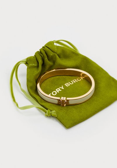 Brățară din metal de culoare aurie, cu un accent din email alb, având un detaliu de închidere, așezată pe un pachet din țesătură verde cu logo.