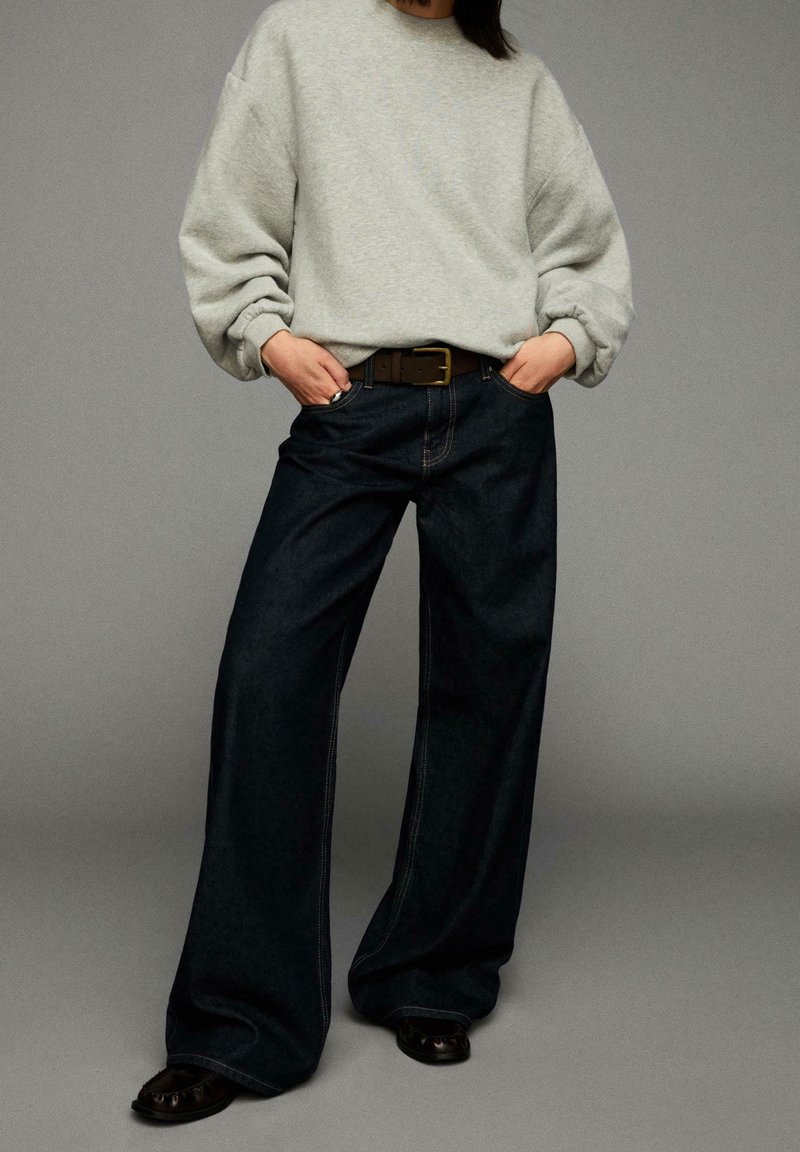 Personne portant un sweat-shirt oversize gris clair rentré dans un jean large foncé avec une ceinture marron et des chaussures noires, debout les mains dans les poches.