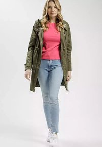 Oliwkowy parka z regulowanymi sznurkami, różowa koszula, jasnoniebieskie dopasowane dżinsy i białe trampki. Gładki materiał, warstwowy wygląd i kaptur.