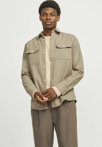 Chemise marron clair avec deux poches avant, fermeture à boutons et manches longues, superposée à un pull beige et assortie à un pantalon beige.
