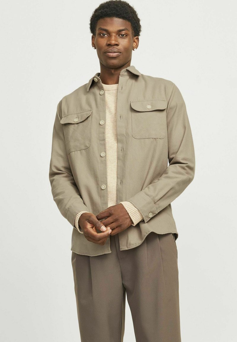 Chemise marron clair avec deux poches avant, fermeture à boutons et manches longues, superposée à un pull beige et assortie à un pantalon beige.