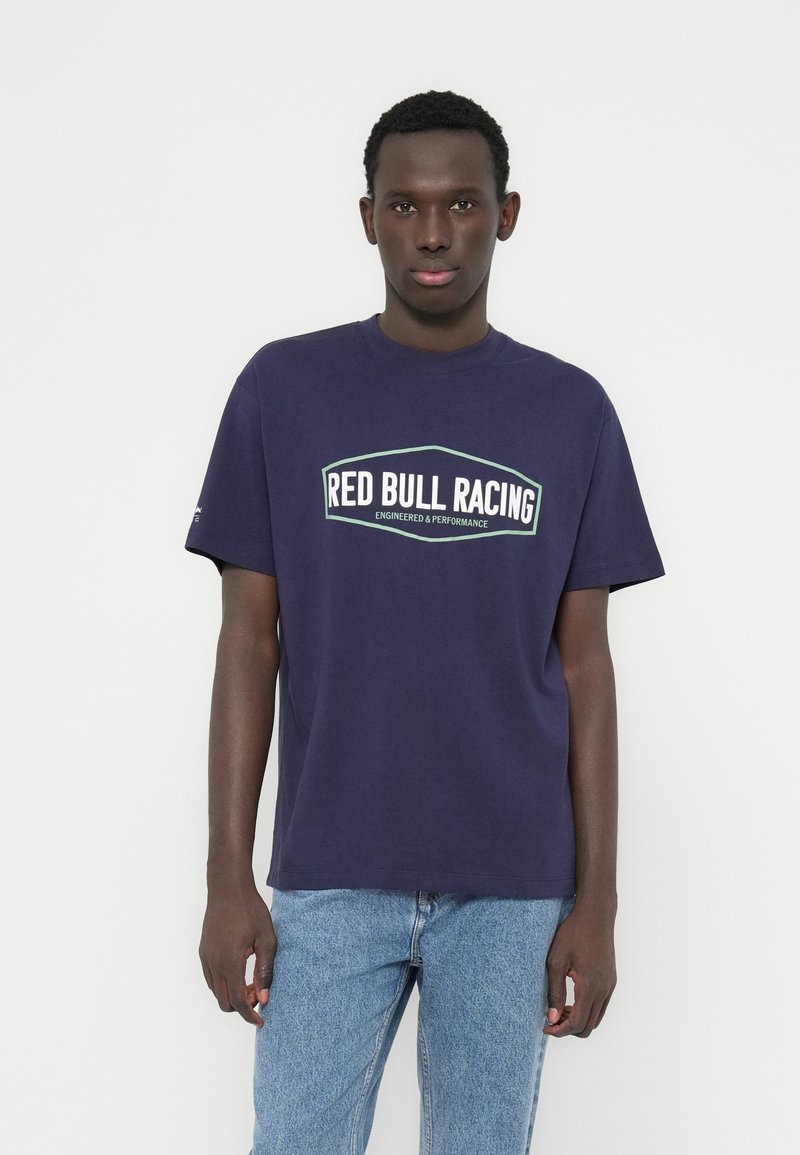 Μπλούζα από βαμβάκι σε ναυτικό μπλε χρώμα με λευκό και πράσινο λογότυπο "RED BULL RACING". Λαιμόκοψη, κοντά μανίκια, άνετη γραμμή. Συνδυασμένη με ανοιχτό μπλε τζιν.