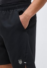 Κοντινό πλάνο ενός ατόμου που φοράει μαύρα αθλητικά σορτς K-Swiss με ελαστική μέση, με το χέρι να ακουμπά στην δεξιά τσέπη.