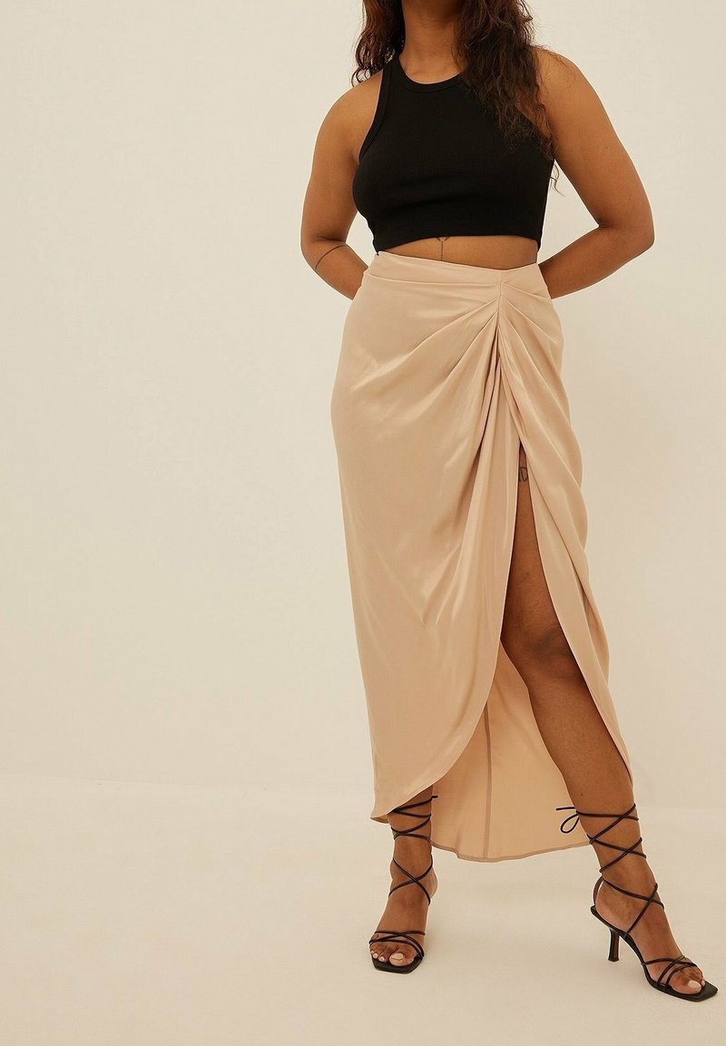 Femme portant un top court noir sans manches, une jupe beige froncée avec fente à la cuisse, et des sandales noires à talons avec des brides, debout sur un fond uni.
