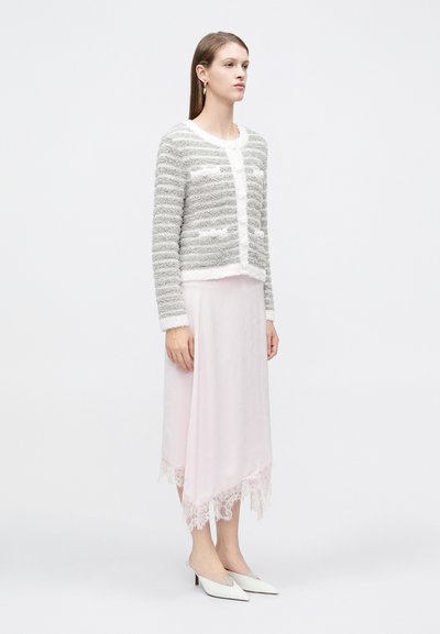 Γυναίκα στέκεται φορώντας γκρι και λευκή ριγέ πλεκτή ζακέτα, φούστα μέσης μήκους σε απαλό ροζ με δαντελωτή λεπτομέρεια και λευκά μυτερά παπούτσια με λουράκι στο πίσω μέρος.