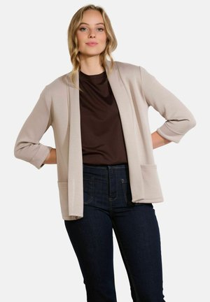 Light beige open-front cardigan with three-quarter sleeves, featuring two front pockets, layered over a dark brown top and dark denim jeans.