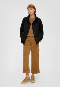 Zwart knopenjas met een kraag, bruine gebreide trui en tan corduroy wijde broek, gecombineerd met beige sneakers.