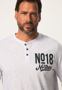 Lys grå henley t-shirt med tre sorte knapper ved udskæringen. Har mørkegrøn tekst: "NO18 Northern Supply by JF1980" på forsiden.