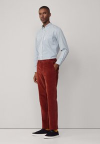 Camisa de botones a rayas en azul claro y blanco, combinada con pantalones de pana en color óxido. Zapatillas oscuras completan el conjunto.