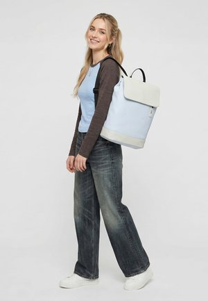 Noor naine, kes kannab helesinist ja halli pika varrukaga särki, tumedaid teksaseid, valgeid tossusid, kannab õlal suurt helesinist seljakotti ja naeratab.