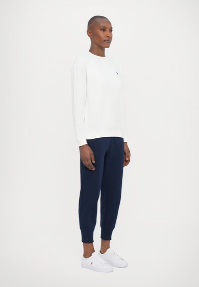 Personne se tenant dans une chemise blanche à manches longues, un pantalon de survêtement bleu marine et des baskets blanches sur un fond clair uni.