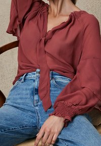 Blusa rossa con fiocco davanti, maniche a sbuffo con polsini arricciati e un tessuto morbido e fluido. Abbinata a jeans a vita alta di colore azzurro chiaro.