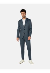 Männer-Sakko in dunkelblauem Stoff mit Reverskragen, zwei Knöpfen und schmal geschnittenen Hosen, kombiniert mit einem einfachen weißen T-Shirt und weißen Sneakern.