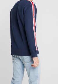 Sweatshirt bleu marine avec des rayures verticales rouges et blanches sur les manches, fabriqué en tissu mélangé de coton, avec un col rond et des poignets côtelés.