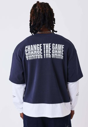 Personne avec des dreadlocks foncés portant une chemise bleu marine sur un t-shirt blanc à manches longues, avec "CHANGE THE GAME" imprimé deux fois dans le dos.