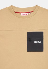 Šviesiai pilkas trumpo rankovio marškinėliai su juodu kišenės pleistru, kuriame yra užtrauktukas ir baltas "HUGO" logotipas. Minkšta medžiaga su lygiu paviršiumi.