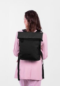 Žena v černém batohu s rolovacím vrškem a zipovými kapsami, oblečená v růžovém obleku, stojí zády k jednoduchému bílému pozadí.