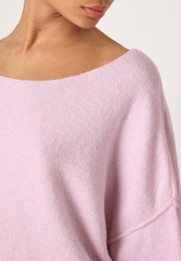 Artimas asmeninės švelnios, šviesiai rožinės plačios apykaklės megztinio vaizdas, rodantis iškirptę, pečių ir dalį veido.