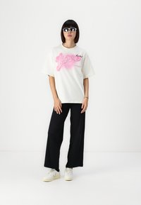 Λευκό oversized t-shirt με ροζ γραφικό σχέδιο, συνδυασμένο με χαλαρές μαύρες παντελόνες και λευκά αθλητικά παπούτσια. Απλό σχέδιο και χαλαρή εφαρμογή.